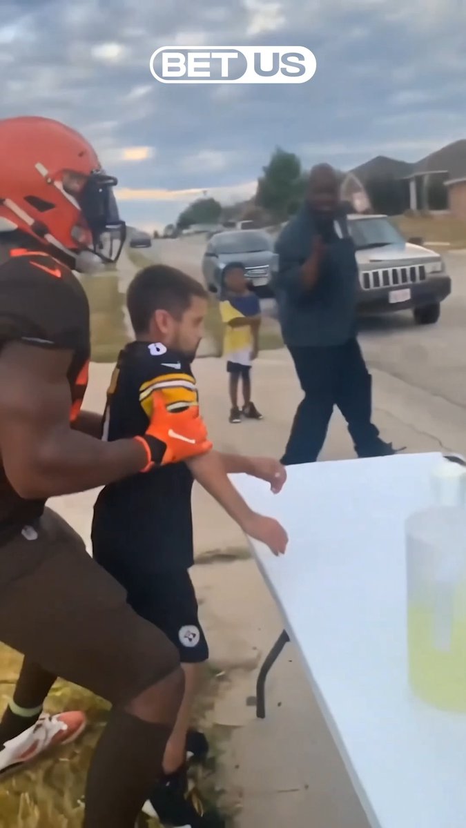 A scene showing an adult in football gear grabbing a young child wearing a black and orange jersey on a suburban street, with a "BET US" logo overlaid at the top.