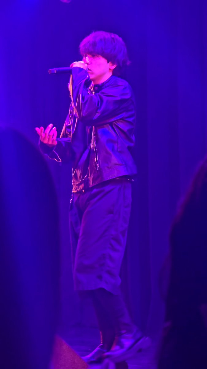 A young man performing on stage under dramatic purple lighting, holding a microphone and wearing a dark jacket.