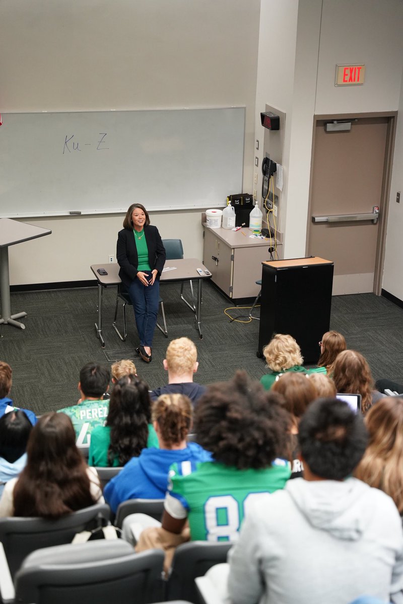 A woman speaks to a seated audience in a classroom. A whiteboard, tables, a podium, and a door are visible; there is text on the board, door sign, and some audience members' clothing.