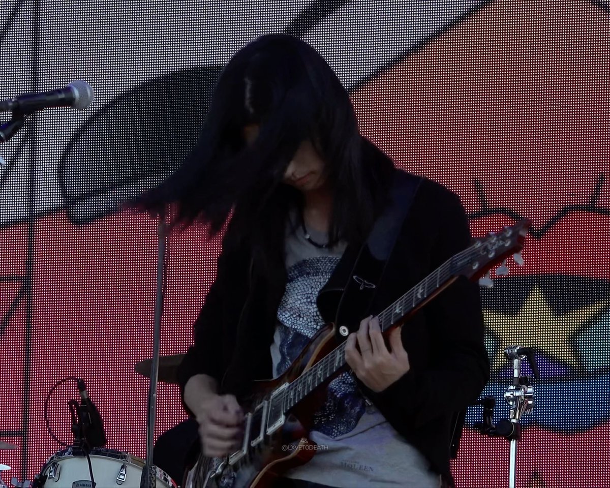A person with long dark hair playing an electric guitar on stage in front of a pixelated LED screen backdrop.