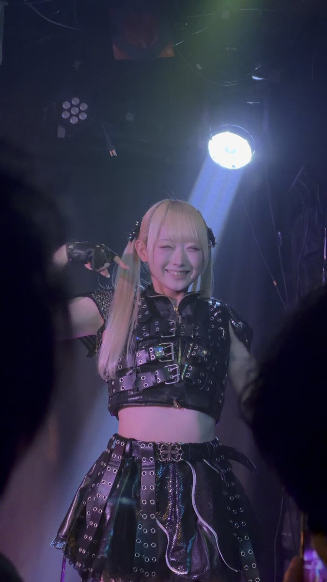 A blonde performer on stage in an edgy black leather harness-style crop top and studded skirt smiles at the camera while making a hand gesture, illuminated by a spotlight.