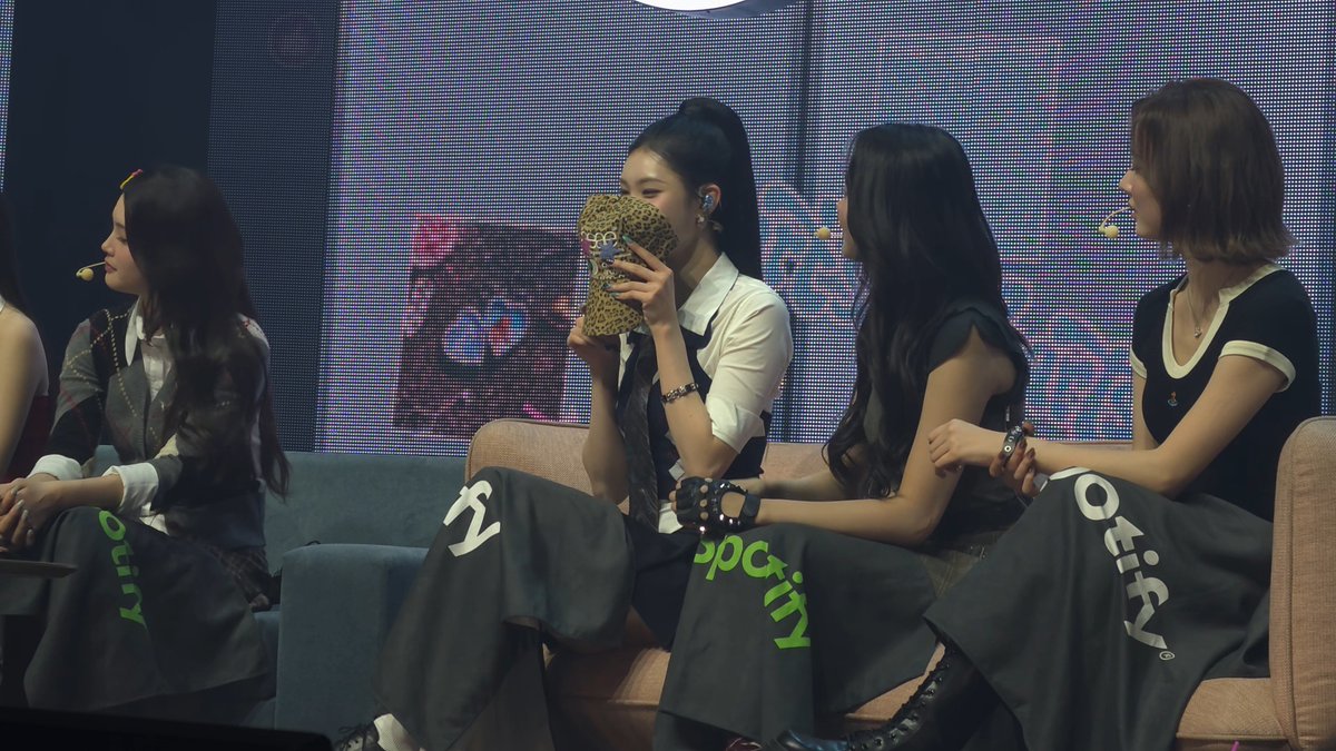 Four young women, apparently K-pop performers, seated on stage during what looks like a fan event; one covers her mouth with a leopard-print phone case while the others wear branded jackets/skirts with 'ROCKIN''.