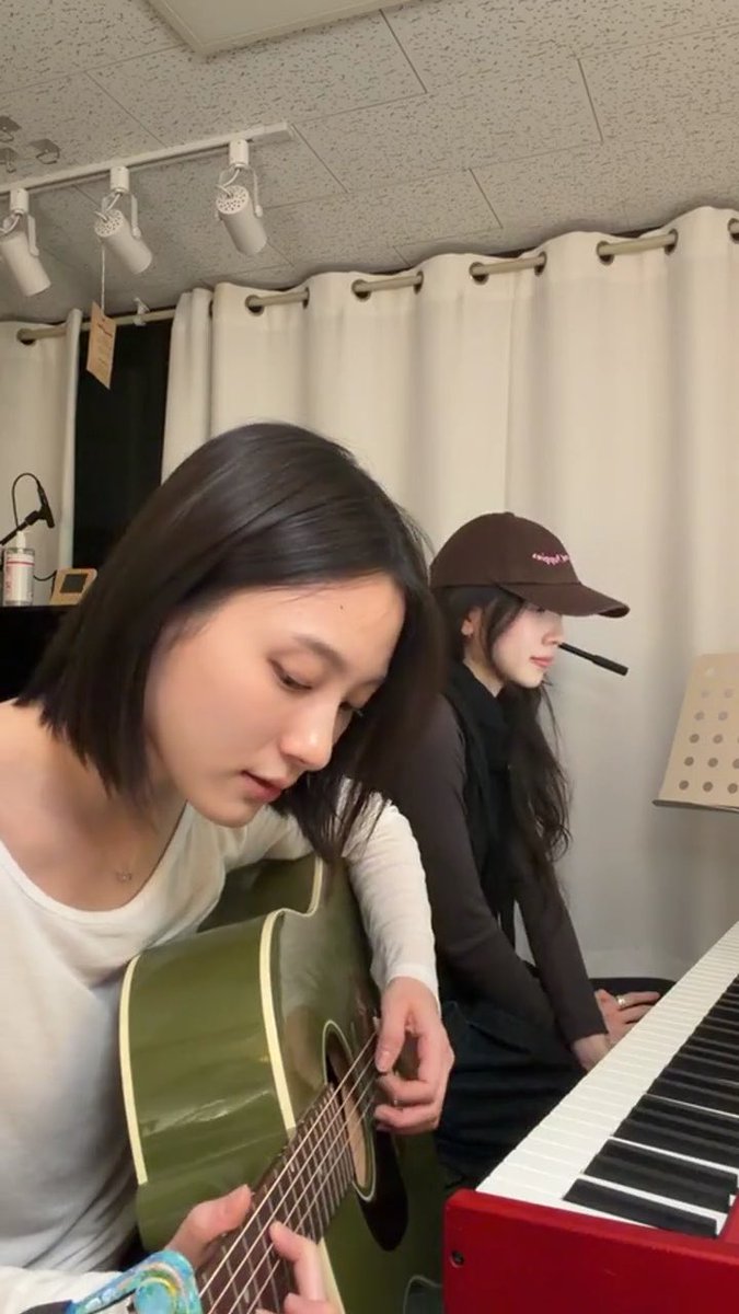 Two young women in a room with white curtains; one in a white shirt plays an acoustic guitar while the other, wearing a brown cap and dark shirt, sits at a red keyboard with a microphone.