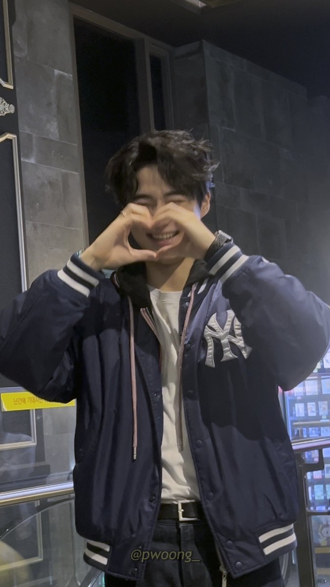 A photo of a smiling young man with dark hair wearing a navy blue NY Yankees varsity jacket, making a heart shape with his hands in front of his face.