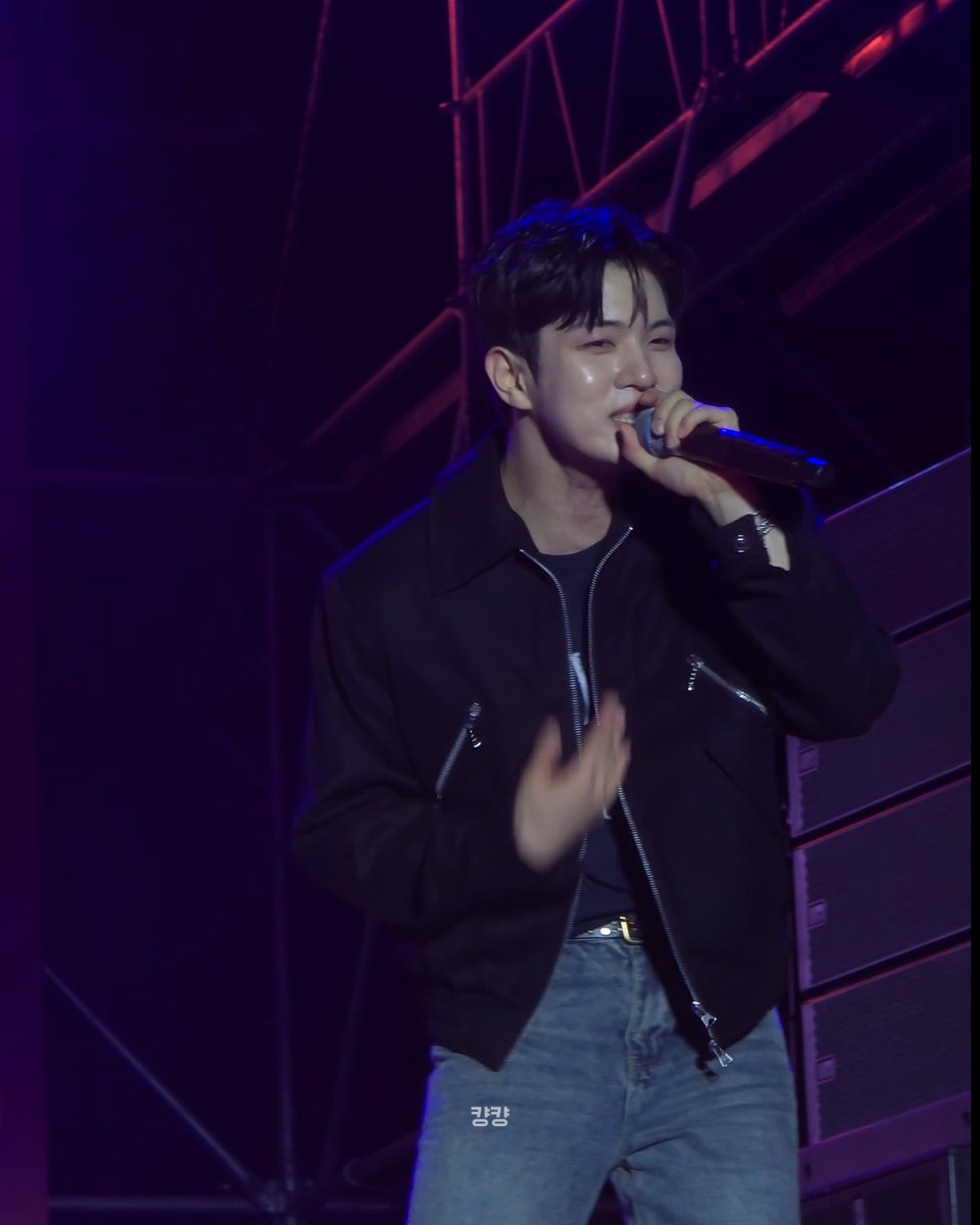 A Korean male performer (K-pop idol) singing into a microphone on a stage with purple/blue lighting, wearing a black zip-up jacket and blue jeans.