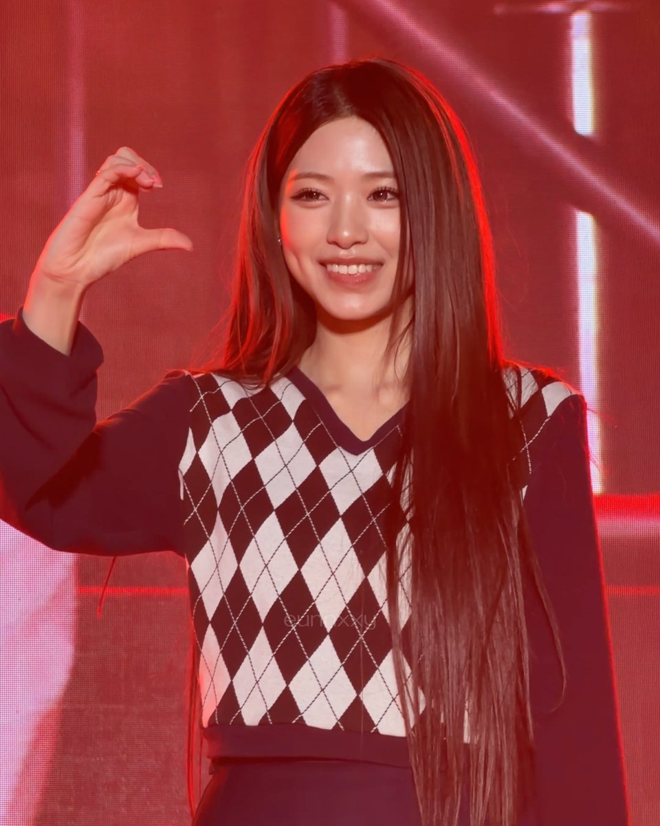 A young woman with long dark hair smiling and making a hand gesture on a stage with a red-lit background, wearing a black and white argyle sweater vest.