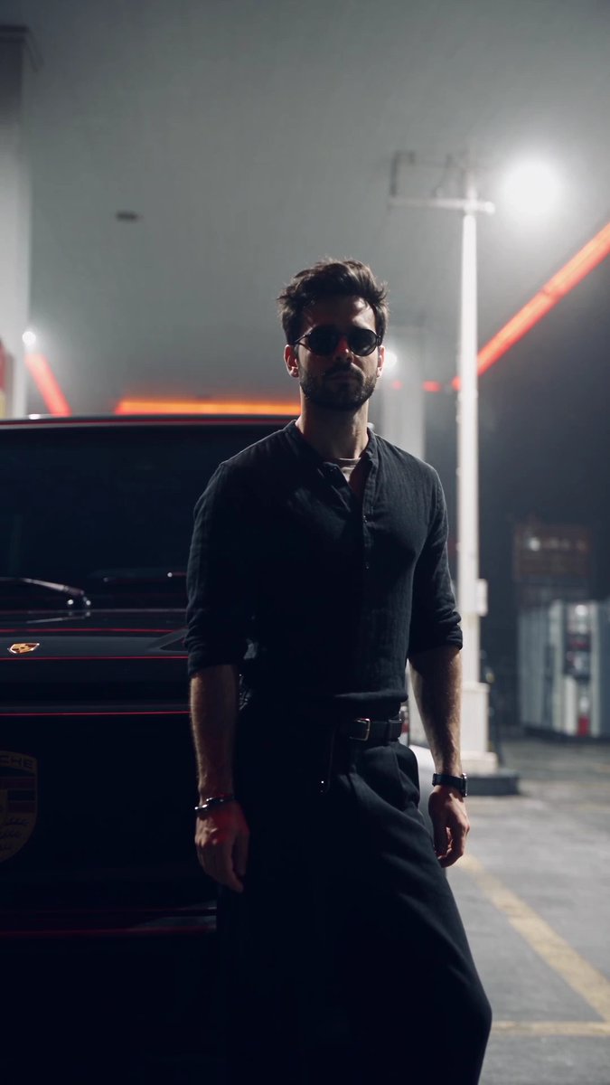 A man in dark sunglasses and a black shirt leans against a dark Porsche with red accents at night, with street lights and power lines visible in the background.