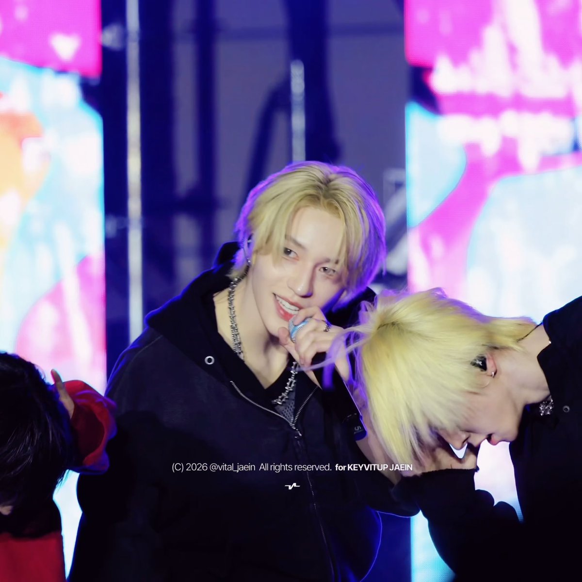 A performer with blonde hair in a black hoodie smiles into a microphone on a colorful stage, with another blonde-haired performer bent over next to him and '(C) 2026 @vital_jaein All rights reserved. for KEYVITUP JAEIN' watermarked across the image.