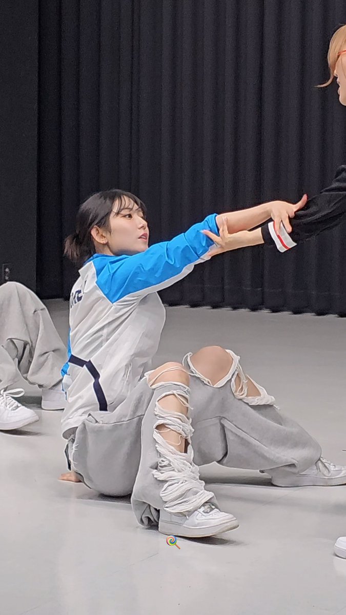 A young woman in a blue and white jacket and ripped gray sweatpants sits on the floor in a dance studio, extending her arm to hold hands with another person standing across from her.
