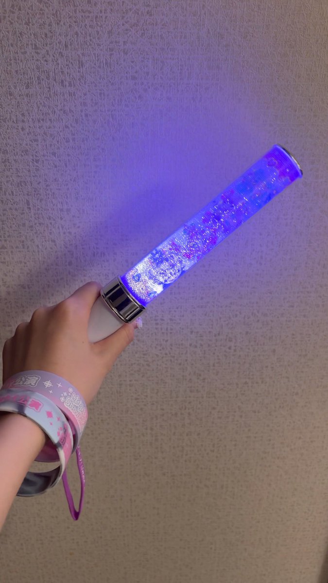 A hand wearing a pink "Wonderful Precure" wristband holds up a glowing lavender and blue glittery pen light against a beige wall, ready for the #TannPriLive performance.