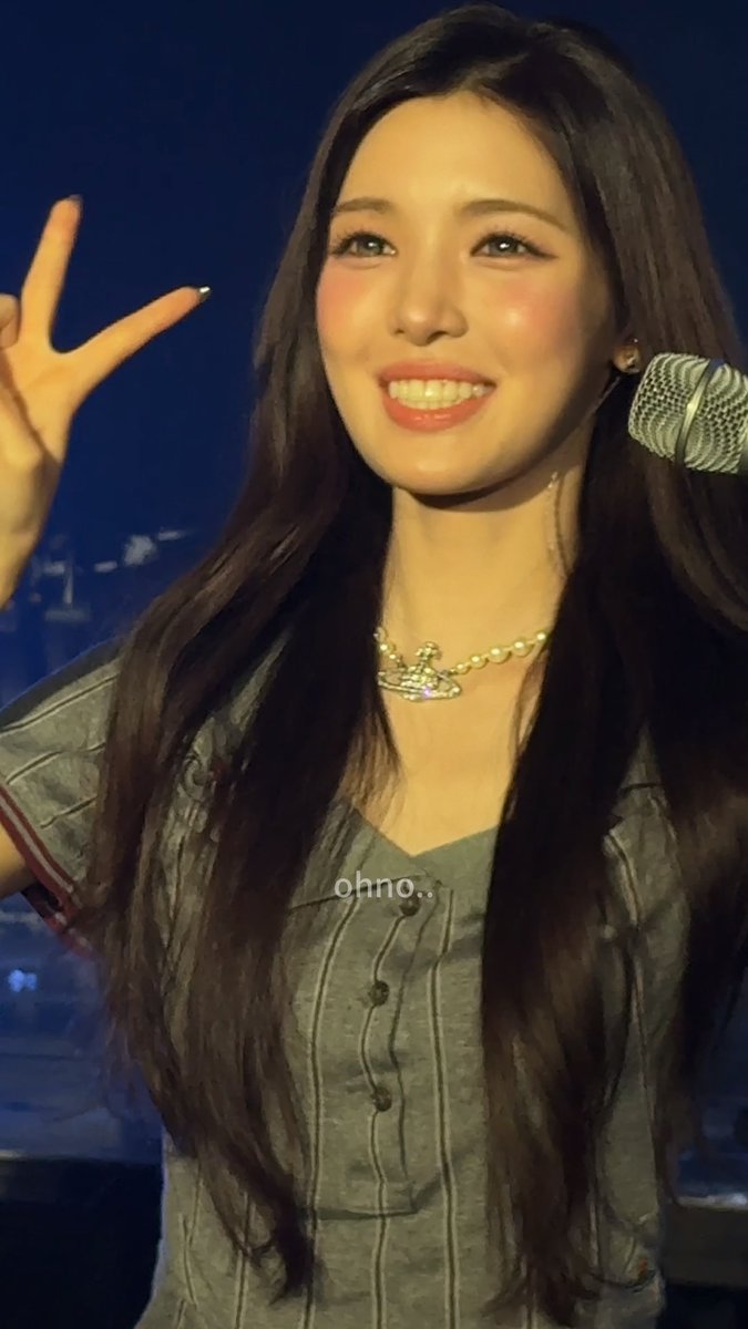 A young woman smiles brightly and flashes a peace sign while standing in front of a microphone, her long dark hair flowing around a gray top and pearl Vivienne Westwood choker, with the text "ohno.." overlaid on the image—capturing the heart-flustered reaction the post describes.