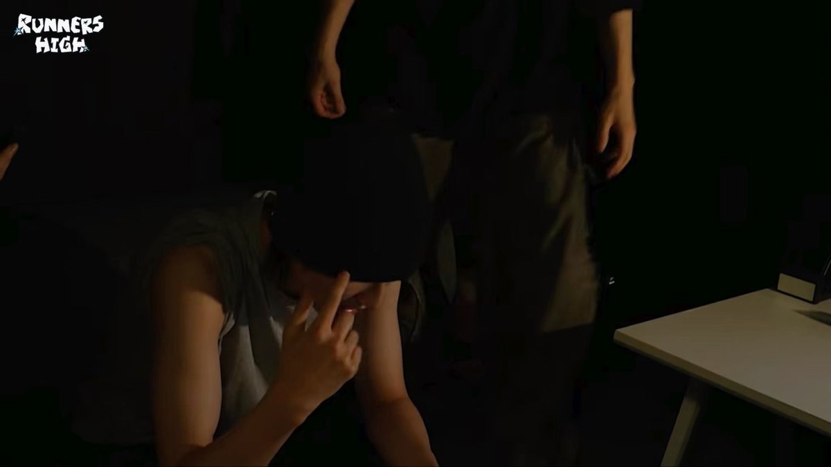 A dark scene from a video or game titled 'RUNNERS HIGH', showing a person sitting at a desk covering their face while seemingly being approached by another figure.