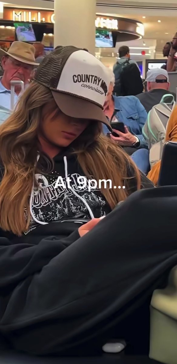 Ella Langley sits in an airport waiting area wearing a "Country Music Association" trucker hat and a black graphic hoodie, looking down at her phone while surrounded by fellow travelers. Text overlay reads "At 1:19pm…" as she waits during her airport delay.