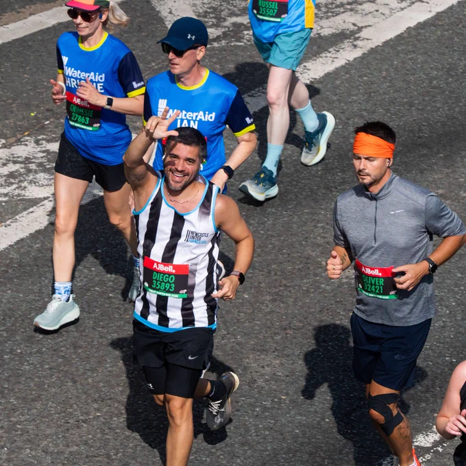 Group of runners in a marathon, with some wearing "WaterAid" shirts. A participant in a black and white striped shirt is smiling and waving at the camera. The race number and branding "ABell" are visible on event bibs.
