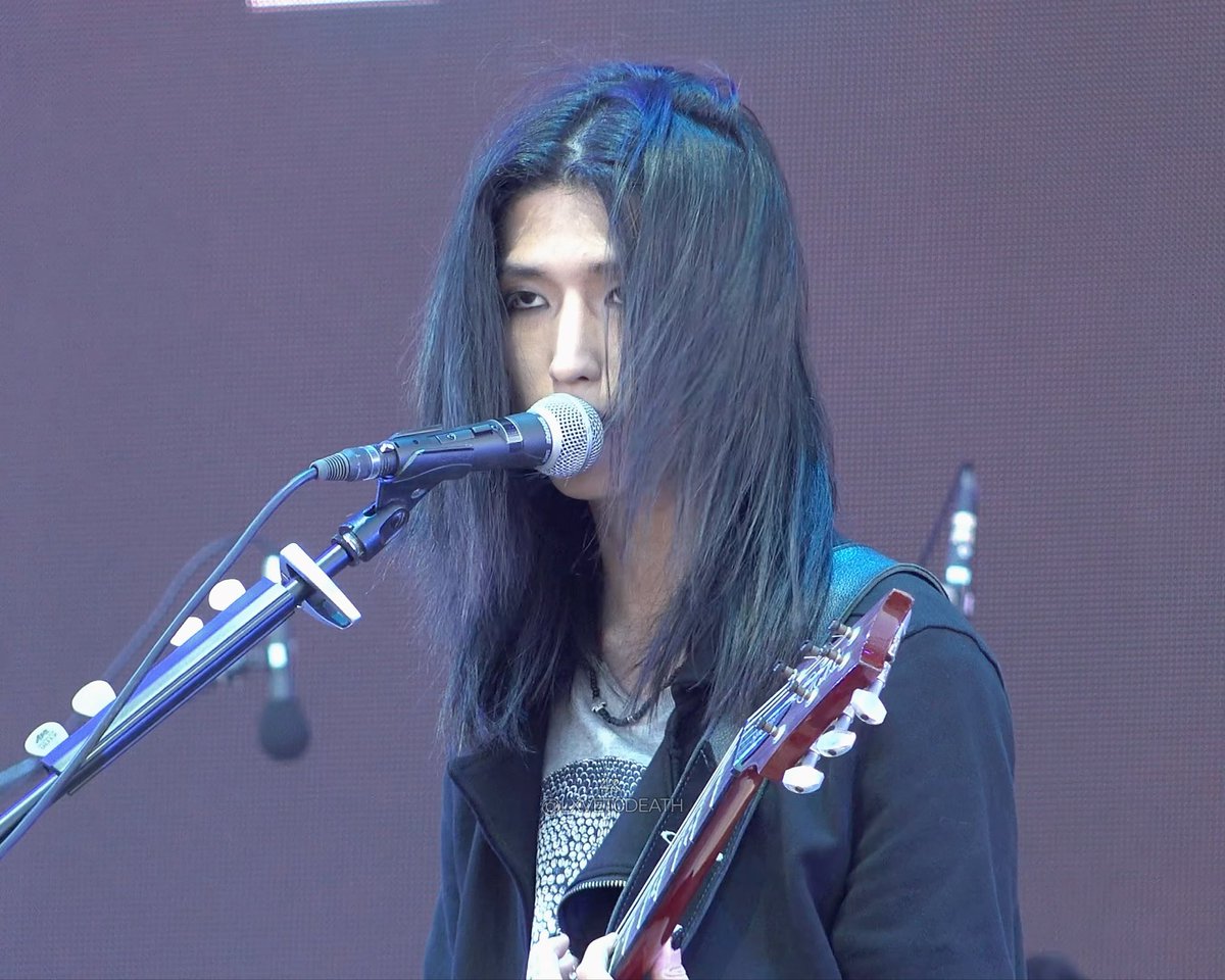 A musician with long dark hair performing on stage, singing into a microphone while playing an electric guitar.