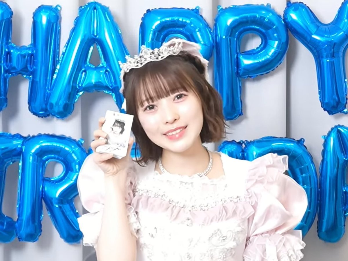 A young woman wearing a tiara and pink frilly dress smiles while holding a small photograph, with large blue 'HAPPY BIRTHDAY' balloons behind her.