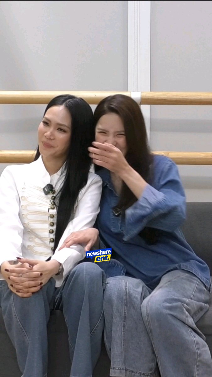 Two women sitting closely on a couch; the woman on the left wears a white shirt, while the woman on the right wears a denim shirt and is laughing while covering her mouth. A "news here ent." watermark is visible.