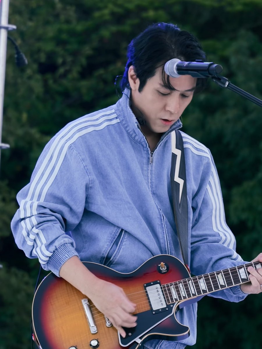 A young man with dark hair plays a sunburst electric guitar while singing into a microphone, wearing a light blue Adidas track jacket with white stripes.