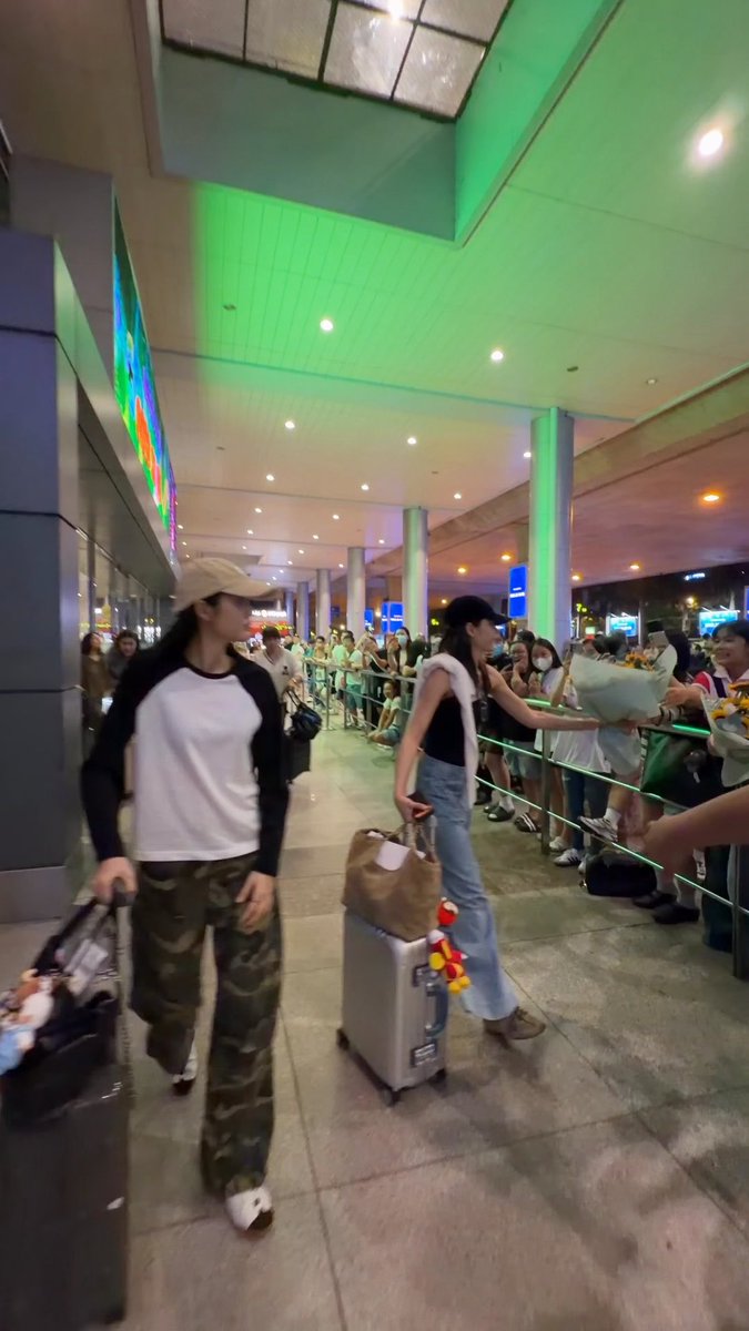 Two women with luggage walk past a crowd of fans holding bouquets of flowers behind a green rope barrier in what appears to be an airport arrival area.