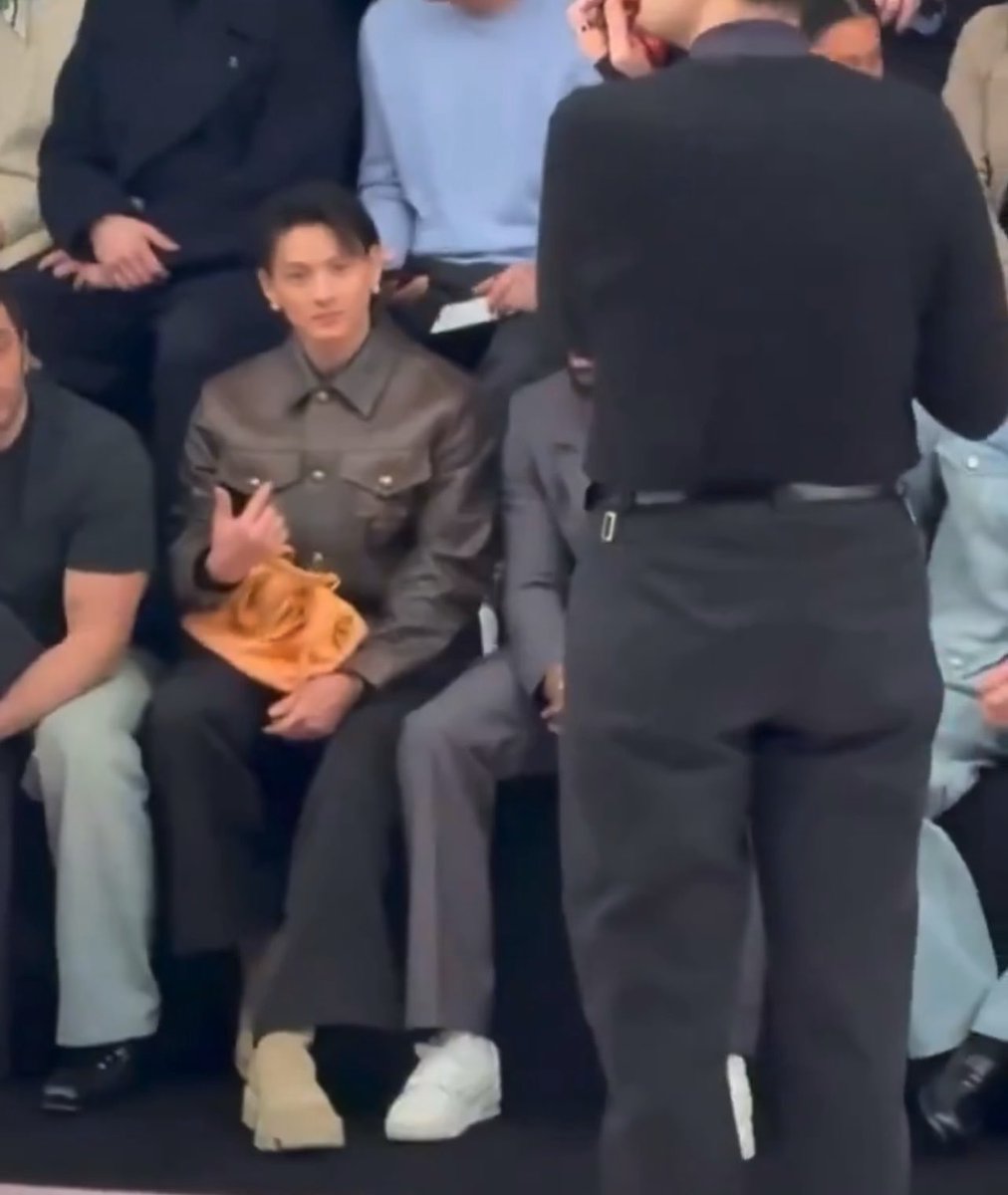 Hirano Sho sits in the front row of what appears to be a fashion show, wearing a brown leather jacket and dark pants while making a peace sign, dressed similarly to those seated around him. The image captures the cute and somewhat awkward moment the post describes, showing him alone in the audience until he noticed someone familiar—matching the caption's playful narrative of finding brief comfort in a foreign setting.