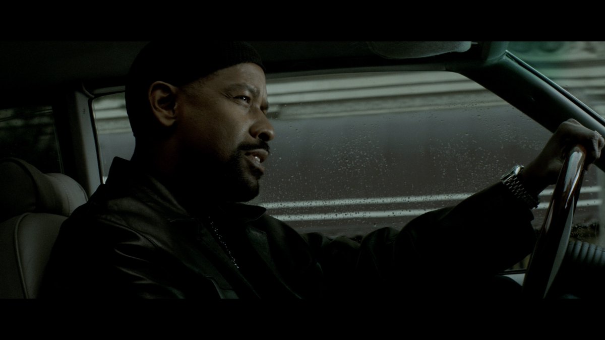 A man in a black cap and leather jacket grips a steering wheel while driving in the rain, looking intently ahead. The image is a scene from the film "Training Day," which is being promoted for a return engagement in Dolby Cinema at AMC Theatres.