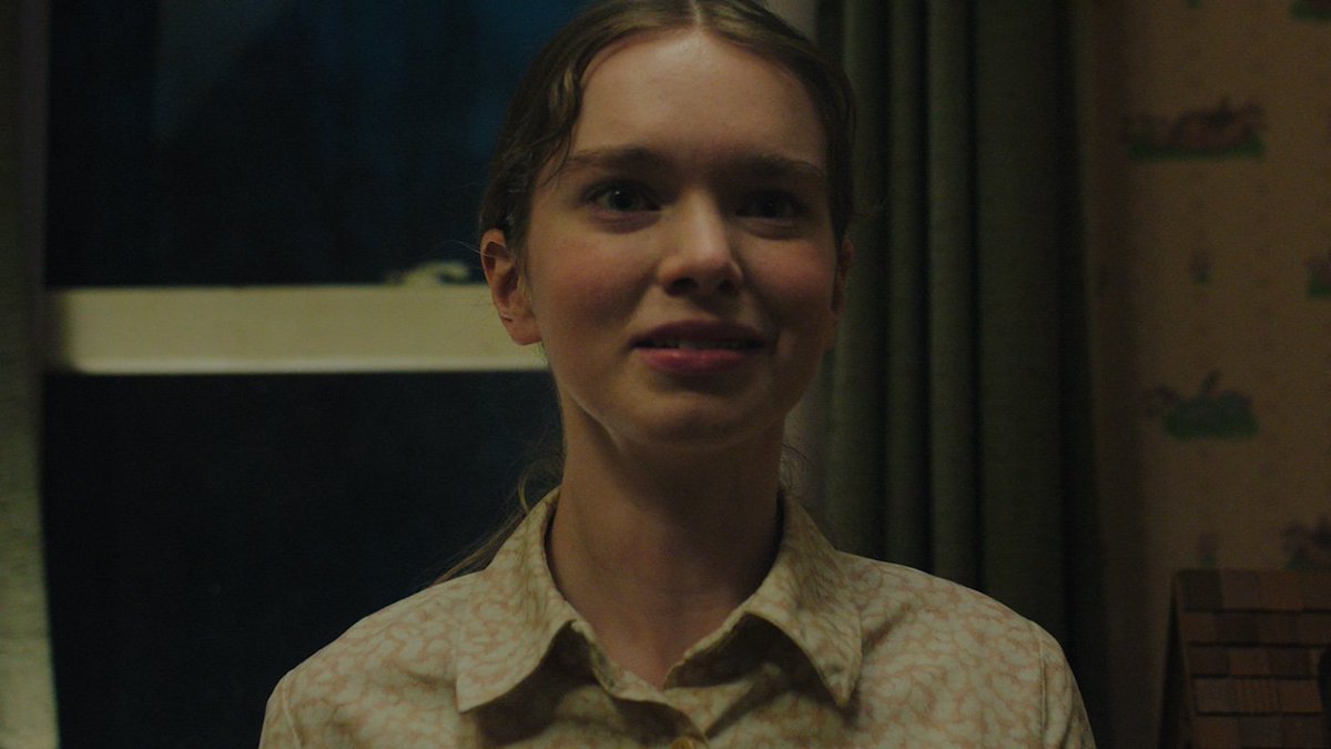 A young woman with light brown hair pulled back looks forward with a subtle expression in a dimly lit room with patterned wallpaper, wearing a beige patterned collared shirt.