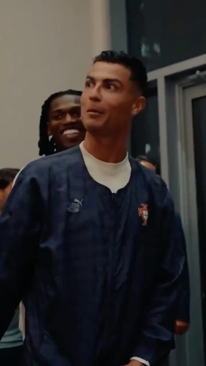 A candid shot of soccer star Cristiano Ronaldo wearing a navy Portugal national team tracksuit jacket with the Puma and team crests, looking to the side while another player smiles behind him.