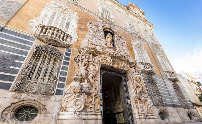 An ornate Baroque-style building facade with intricate stone carvings, decorative balconies, and a central archway featuring sculptural figures.