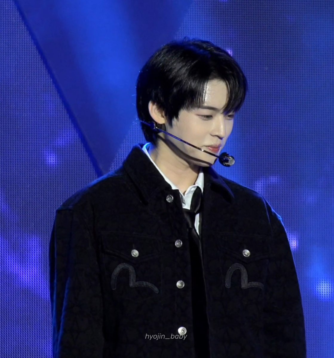 A young man with dark hair performing on stage with a headset microphone, wearing a black jacket with decorative embroidery over a white shirt. The photo is watermarked 'hyojin_baby' against a blue stage backdrop.
