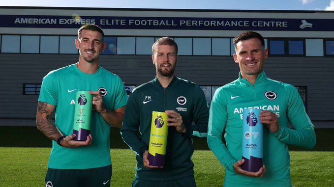 Three men in green Brighton & Hove Albion training gear posing on a football pitch at the American Express Elite Football Performance Centre, each holding a cylindrical Premier League award.