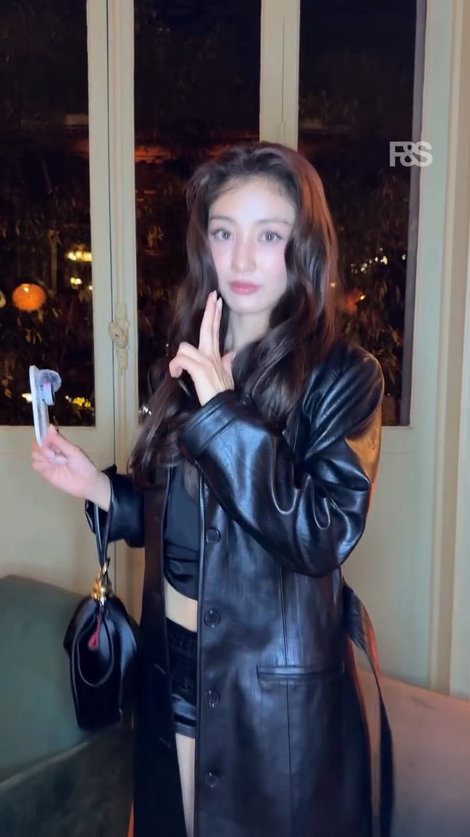 Jihyo, the K-pop star, poses indoors in a sleek black leather coat with long wavy hair, holding a small item while making a peace sign, fitting the post's caption of her appearance at the Christian Louboutin SS27 show. The "F&S" watermark is visible in the upper right corner of the image.