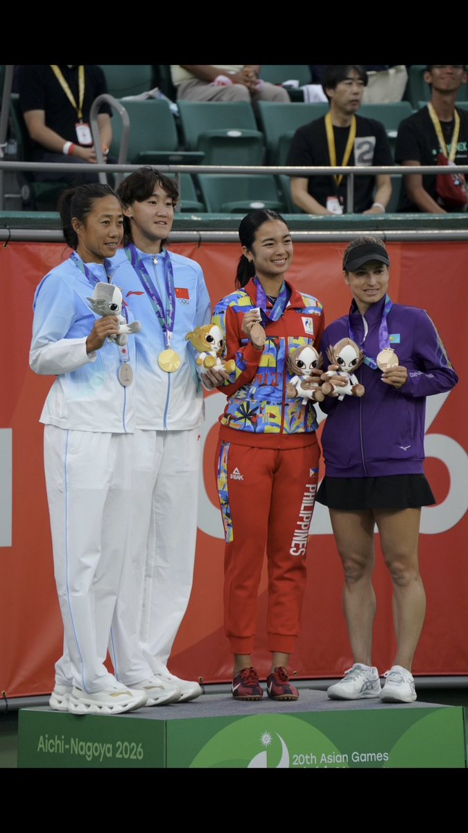 Four female athletes stand on a podium at the 20th Asian Games (Aichi-Nagoya 2026), proudly displaying their medals and mascot plushies, with athletes wearing uniforms representing China, the Philippines, and Kazakhstan.