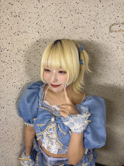 A cosplayer with blonde hair and blue hair clips posing in a blue and white frilly dress against a textured wall.