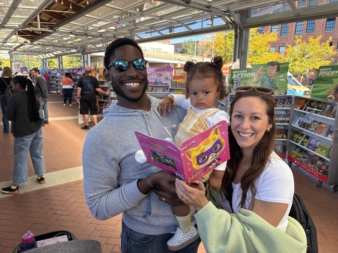 Two adults, one with a young child in their arms, are smiling at the cameras while holding a book.
