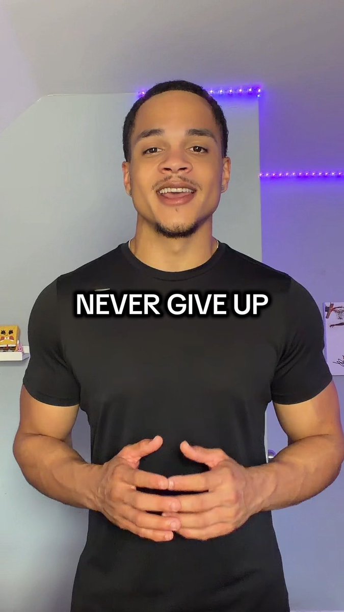 A young man in a black t-shirt smiles at the camera with his hands clasped, with the text 'NEVER GIVE UP' displayed across his chest.