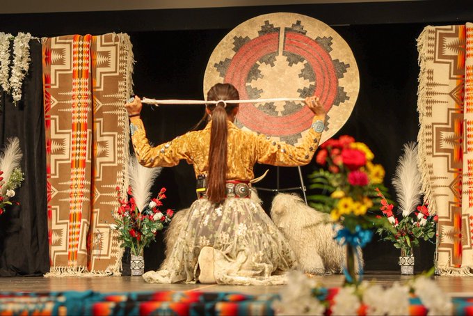 A Native American dancer viewed from behind, wearing traditional attire and holding a long stick horizontally, with a large circular patterned artwork displayed on the black wall behind them.