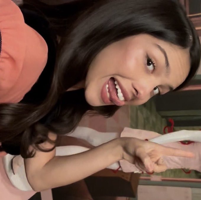 A woman with long dark hair in a pink top playfully poses sideways, flashing a peace sign with her mouth open in an animated expression.