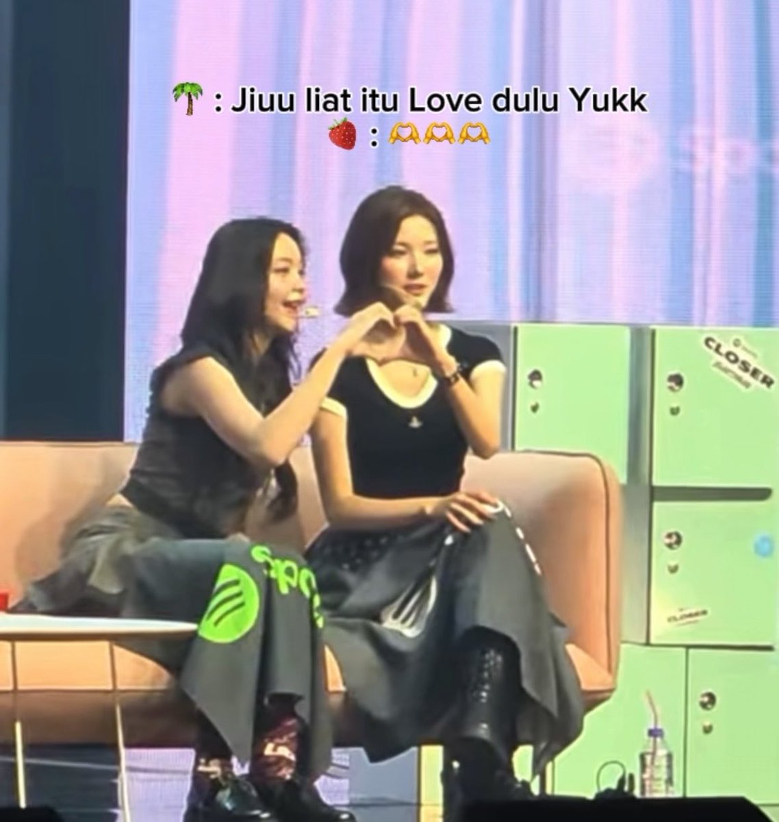 Two women sit on a peach-colored couch on a stage, with the one on the left forming a heart shape with the other woman's arms; overlaid text reads '🌴 : Jiuu liat itu Love dulu Yukk' and '🍓 : 🫶🫶🫶'.