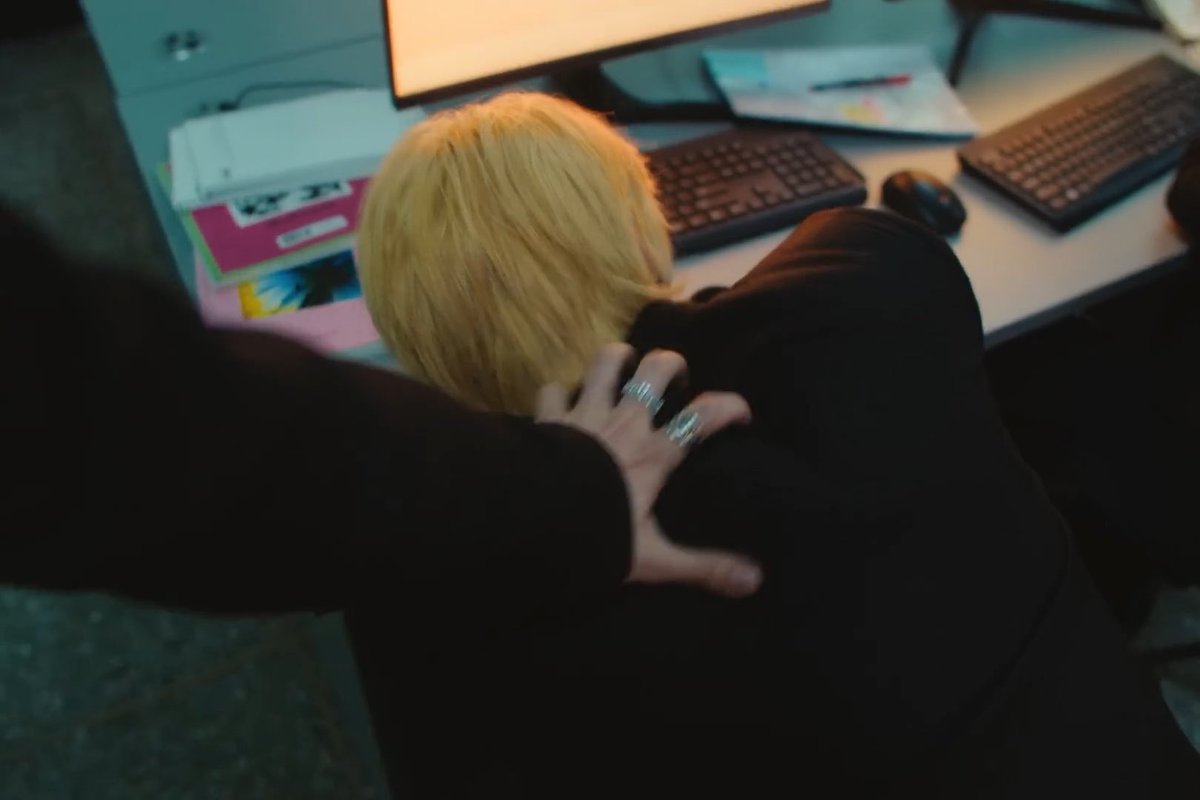 Photograph taken from behind a person with short blonde hair wearing black, showing another person's hand with silver rings resting on their shoulder, set in an office environment with keyboards and a printer visible on a desk.