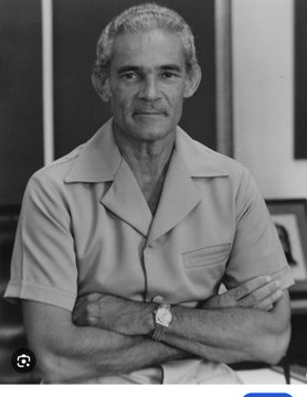 A black-and-white portrait photograph of an older man with gray hair and a mustache, wearing a short-sleeve shirt and a wristwatch, standing with his arms crossed.