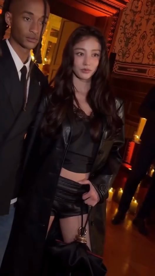 A young woman in a black leather jacket and lace top stands beside a tall man in a suit at what appears to be a formal event with ornate decor.