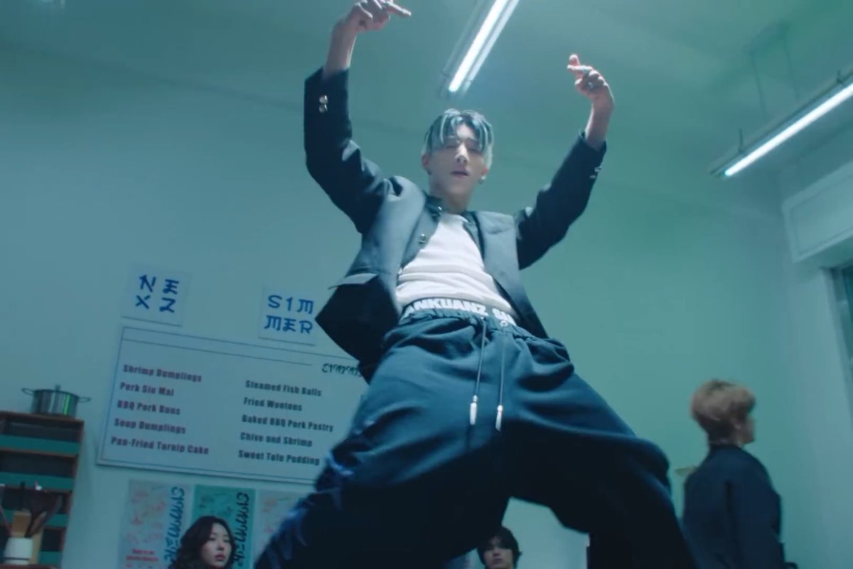 A low-angle shot of a young man in a black jacket and dark pants striking a dynamic pose with raised arms in what appears to be a dance studio or kitchen set.