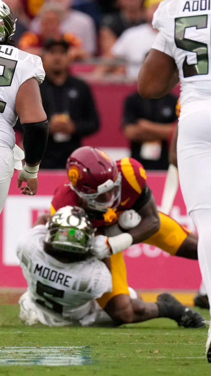 A USC player in a cardinal and gold helmet tackles an Oregon player named "MOORE" (#5), while another Oregon player (#2, partially visible as "ADDI") stands nearby on the field. The image captures a moment of intense physical contact, illustrating the post's theme of controversial and aggressive moments in college football, as Moore appears to be on the ground after a hard hit.
