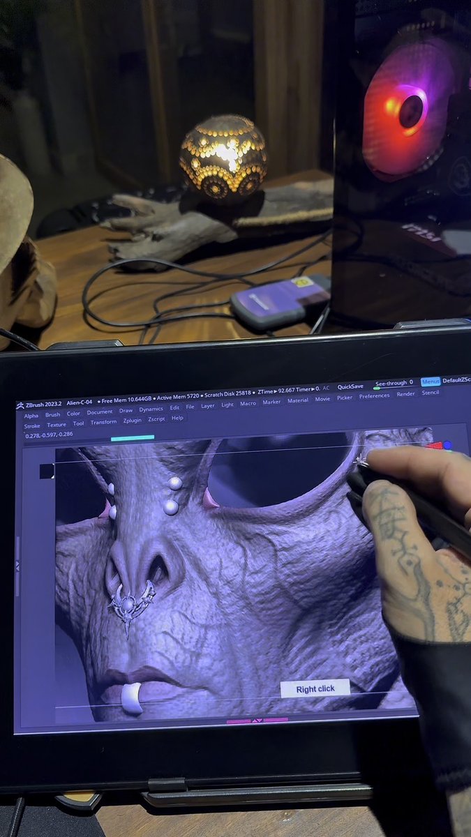 A tattooed hand working on a 3D character model in ZBrush 2023.2 software on a laptop, with an MSI laptop and decorative lamp visible in the background.
