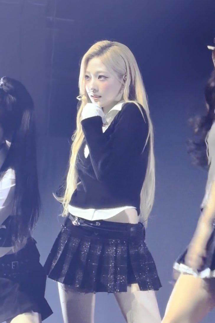 Another stage performance photo of Karina (aespa) in the same black sweater and pleated skirt outfit, looking to the side under stage lighting.