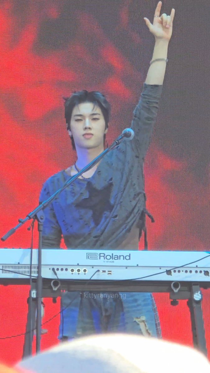 A performer with short dark hair stands behind a Roland keyboard on stage with a red backdrop, wearing a loose black distressed shirt.