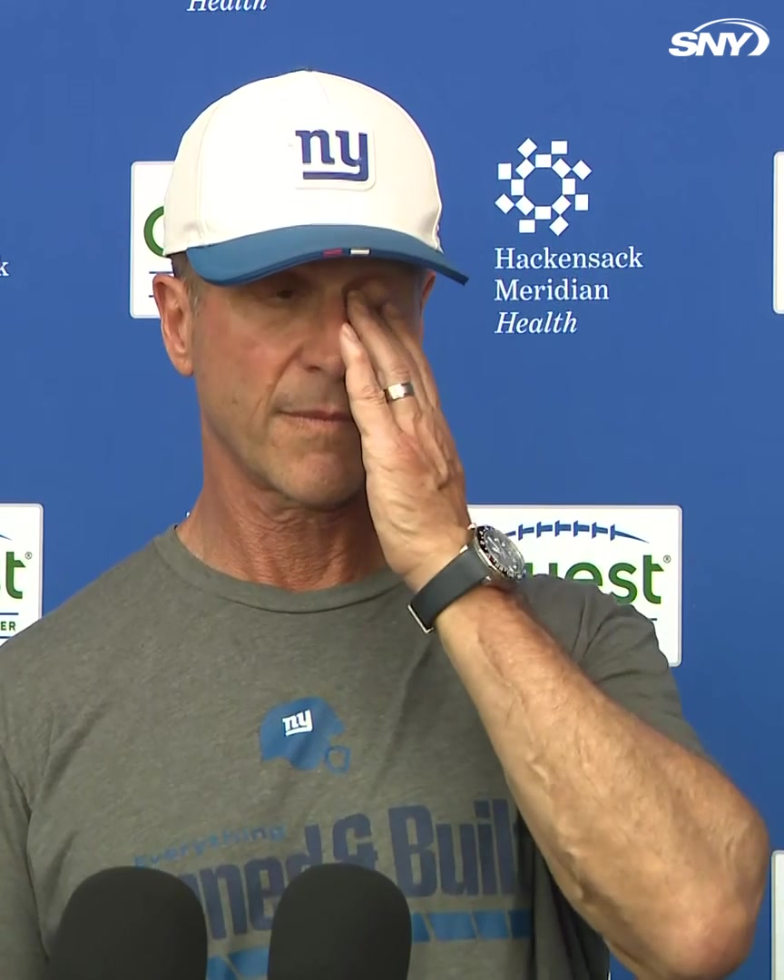 Giants head coach John Harbaugh, wearing a New York Giants cap and shirt, appears emotional during a press conference, rubbing his eye while speaking with reporters about center John Michael Schmitz's absence from practice.