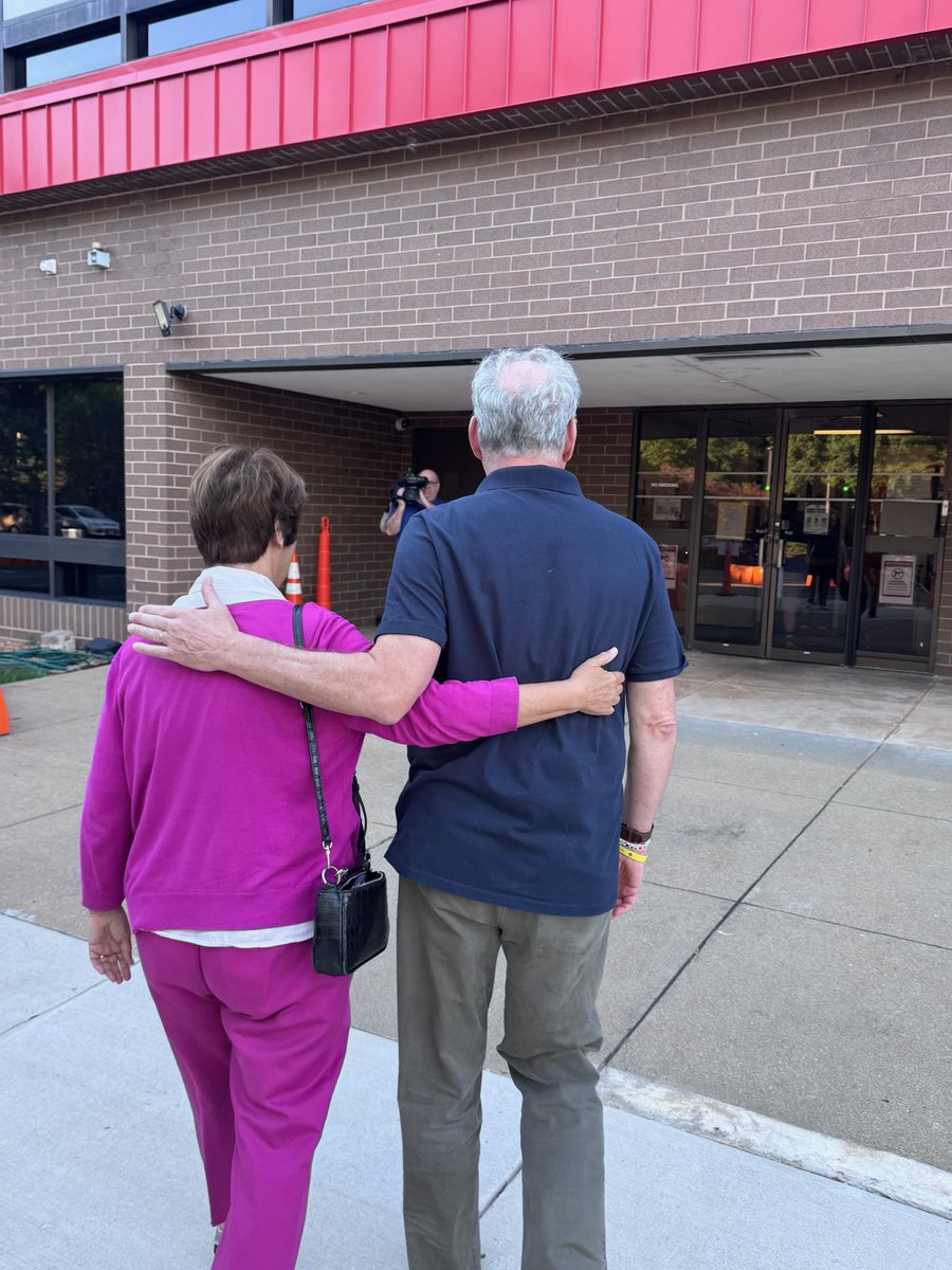 Two older adults walk arm in arm toward the entrance of a brick building. A person holding a camera stands near the doorway, and there is some text on the glass doors.