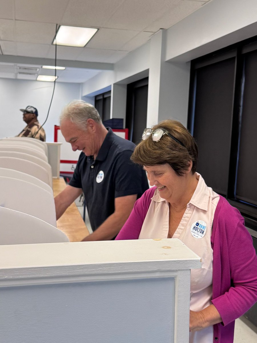 Two people work on or move a large white counter or cabinet in an indoor room, while another person stands in the background. There is text on a badge worn by the woman.