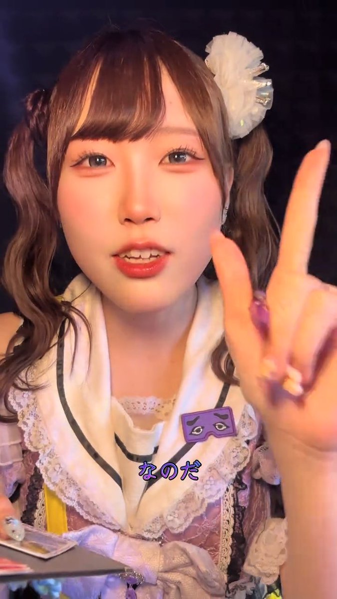 A Japanese idol/performer in a frilly costume with a white flower hair accessory points toward the camera, with Japanese subtitles 'なのだ' displayed.